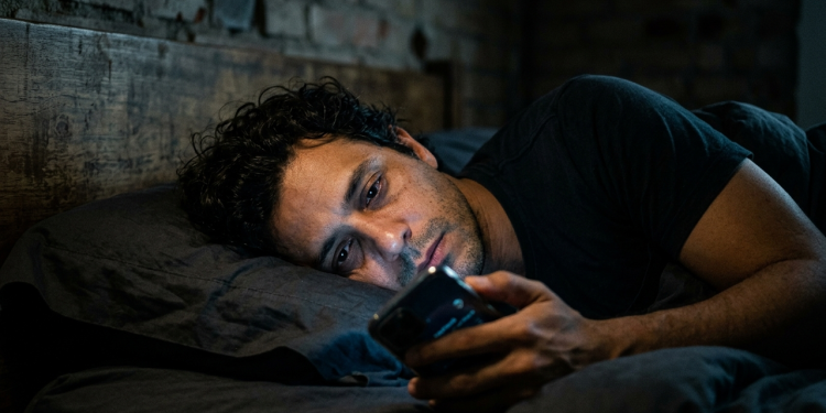 Homem exausto iluminado pela luz azul do celular no escuro.