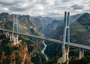 [fotografia editorial de alta qualidade, estilo revista de engenharia civil, infraestrutura e logística, com atmosfera de escala extrema e relevo vencido / Ponte Duge (Beipanjiang Bridge) em destaque absoluto — tabuleiro elevado sobre o cânion profundo entre Guizhou e Yunnan, com torres estaiadas e cabos suspensos, a paisagem montanhosa ao redor, a luz natural criando reflexos e contrastes, transmitindo a sensação de engenharia que venceu o relevo extremo / composição sangrada: a ponte e o cânion ocupam o centro da imagem, com as montanhas, o vale e o céu estendendo-se até as bordas, sem margens ou moldura / profundidade de campo cinematográfica: foco nítido na ponte e nos cabos, com as montanhas e o horizonte suavemente desfocados com bokeh orgânico, sugerindo a escala e a integração com a paisagem / iluminação técnica e dramática: luz natural de dia com nuvens, criando reflexos e contrastes na estrutura metálica e no concreto, com áreas de luz e sombra que destacam a ponte e o vale, transmitindo a sensação de precisão e engenharia, sem artificialidade de estúdio / ângulo chamativo: plano geral com câmera posicionada na altura do vale ou ligeiramente aérea, capturando a ponte em perspectiva, os cabos, o tabuleiro, as montanhas, sugerindo a escala e a travessia extrema / paleta emocional do tema Construção com toque montanhoso: azul-aço na estrutura, cinza-metal nos cabos, branco-gelo nos reflexos, terracota e ocre nas montanhas, verde-selva na paisagem, concreto na estrutura / ambiente contextualizado: Guizhou/Yunnan, China, Ponte Duge, cânion, tabuleiro elevado, cabos estaiados, montanhas, elementos que sugerem a engenharia extrema e a conexão regional, sem cenário encenado artificialmente / sem texto visível, sem pessoas em destaque, sem elementos de stock / atmosfera: engenharia extrema, relevo vencido, a travessia que liga montanhas suspensas no céu / 16:9, composição sangrada sem margens, alta qualidade editorial, fotografia com luz natural e atmosfera de infraestrutura e montanha]