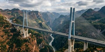 [fotografia editorial de alta qualidade, estilo revista de engenharia civil, infraestrutura e logística, com atmosfera de escala extrema e relevo vencido / Ponte Duge (Beipanjiang Bridge) em destaque absoluto — tabuleiro elevado sobre o cânion profundo entre Guizhou e Yunnan, com torres estaiadas e cabos suspensos, a paisagem montanhosa ao redor, a luz natural criando reflexos e contrastes, transmitindo a sensação de engenharia que venceu o relevo extremo / composição sangrada: a ponte e o cânion ocupam o centro da imagem, com as montanhas, o vale e o céu estendendo-se até as bordas, sem margens ou moldura / profundidade de campo cinematográfica: foco nítido na ponte e nos cabos, com as montanhas e o horizonte suavemente desfocados com bokeh orgânico, sugerindo a escala e a integração com a paisagem / iluminação técnica e dramática: luz natural de dia com nuvens, criando reflexos e contrastes na estrutura metálica e no concreto, com áreas de luz e sombra que destacam a ponte e o vale, transmitindo a sensação de precisão e engenharia, sem artificialidade de estúdio / ângulo chamativo: plano geral com câmera posicionada na altura do vale ou ligeiramente aérea, capturando a ponte em perspectiva, os cabos, o tabuleiro, as montanhas, sugerindo a escala e a travessia extrema / paleta emocional do tema Construção com toque montanhoso: azul-aço na estrutura, cinza-metal nos cabos, branco-gelo nos reflexos, terracota e ocre nas montanhas, verde-selva na paisagem, concreto na estrutura / ambiente contextualizado: Guizhou/Yunnan, China, Ponte Duge, cânion, tabuleiro elevado, cabos estaiados, montanhas, elementos que sugerem a engenharia extrema e a conexão regional, sem cenário encenado artificialmente / sem texto visível, sem pessoas em destaque, sem elementos de stock / atmosfera: engenharia extrema, relevo vencido, a travessia que liga montanhas suspensas no céu / 16:9, composição sangrada sem margens, alta qualidade editorial, fotografia com luz natural e atmosfera de infraestrutura e montanha]